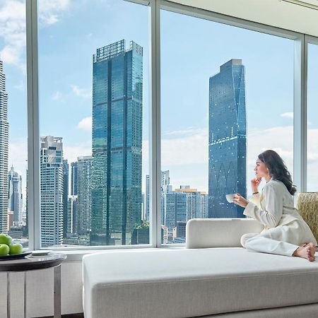 Traders Hotel, קואלה לומפור מראה חיצוני תמונה The photo shows a woman sitting on a comfortable couch, gazing out of a large window. She is dressed in cozy, light-colored clothing and holding a cup, possibly enjoying a beverage. Outside the window, there is a panoramic view of a modern cityscape,
