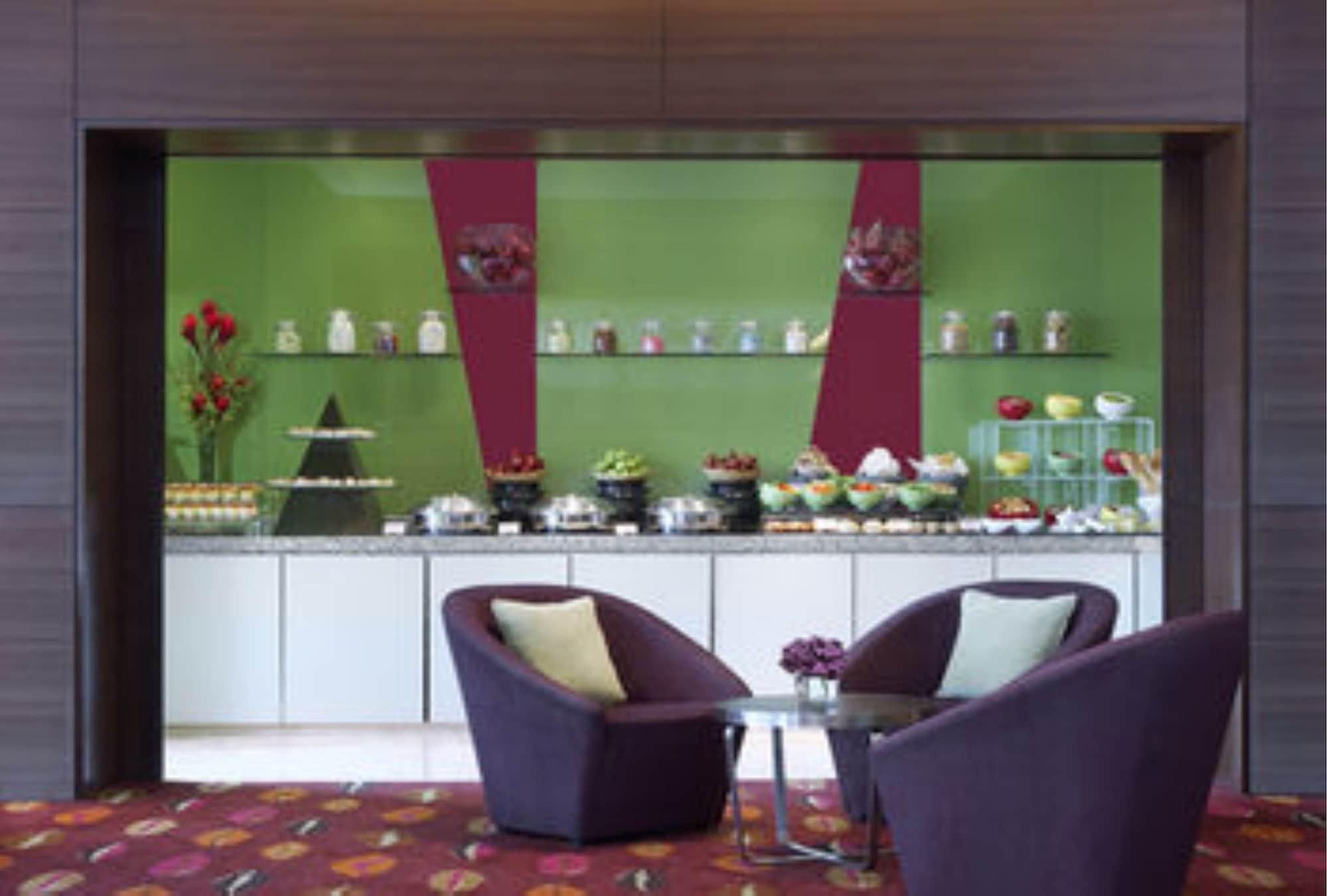 Traders Hotel, קואלה לומפור מראה חיצוני תמונה The photo shows a modern cafe or lounge area featuring two stylish, purple chairs with cushions and a small table between them. Behind the seating area, there is a display counter with an array of colorful desserts and snacks, including cakes, pastri