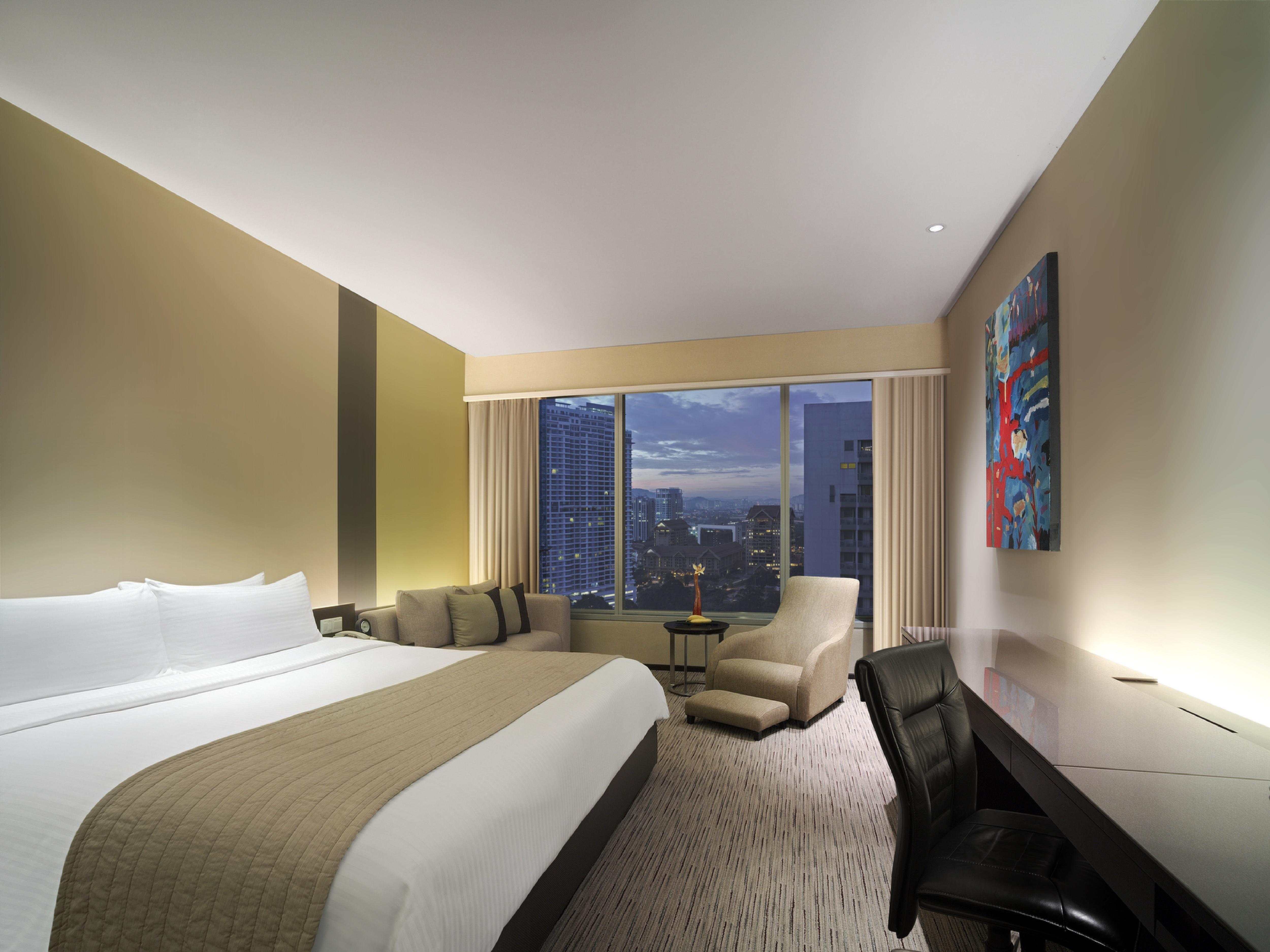 Traders Hotel, קואלה לומפור מראה חיצוני תמונה The photo shows a modern hotel room with a spacious layout. There is a large bed with white linens and a beige headboard, positioned against a wall. To the side, there's a cozy seating area with a couch and a chair. A desk and black office chair are 