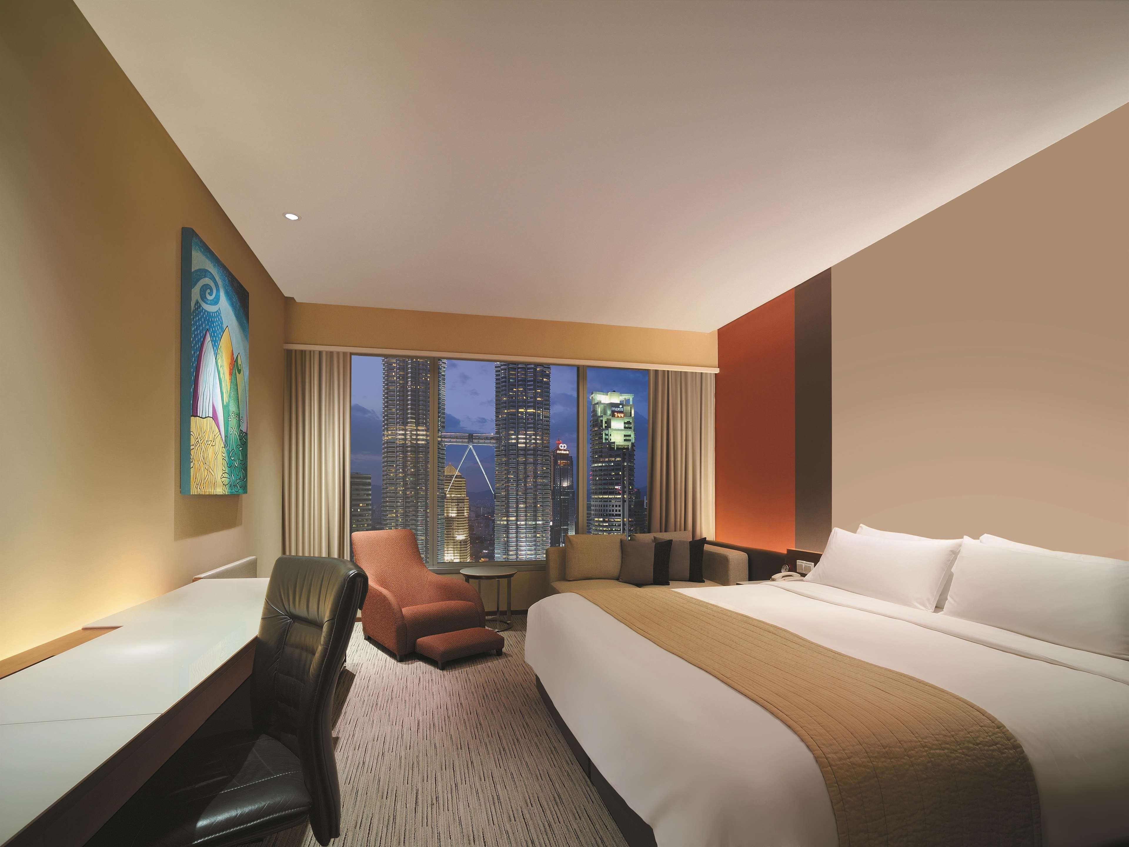 Traders Hotel, קואלה לומפור מראה חיצוני תמונה The photo depicts a modern hotel room featuring a large bed with crisp white linens and a brown throw blanket. There is a comfortable chair and small table beside the bed, as well as a workspace with a desk and an office chair. A large window offers 
