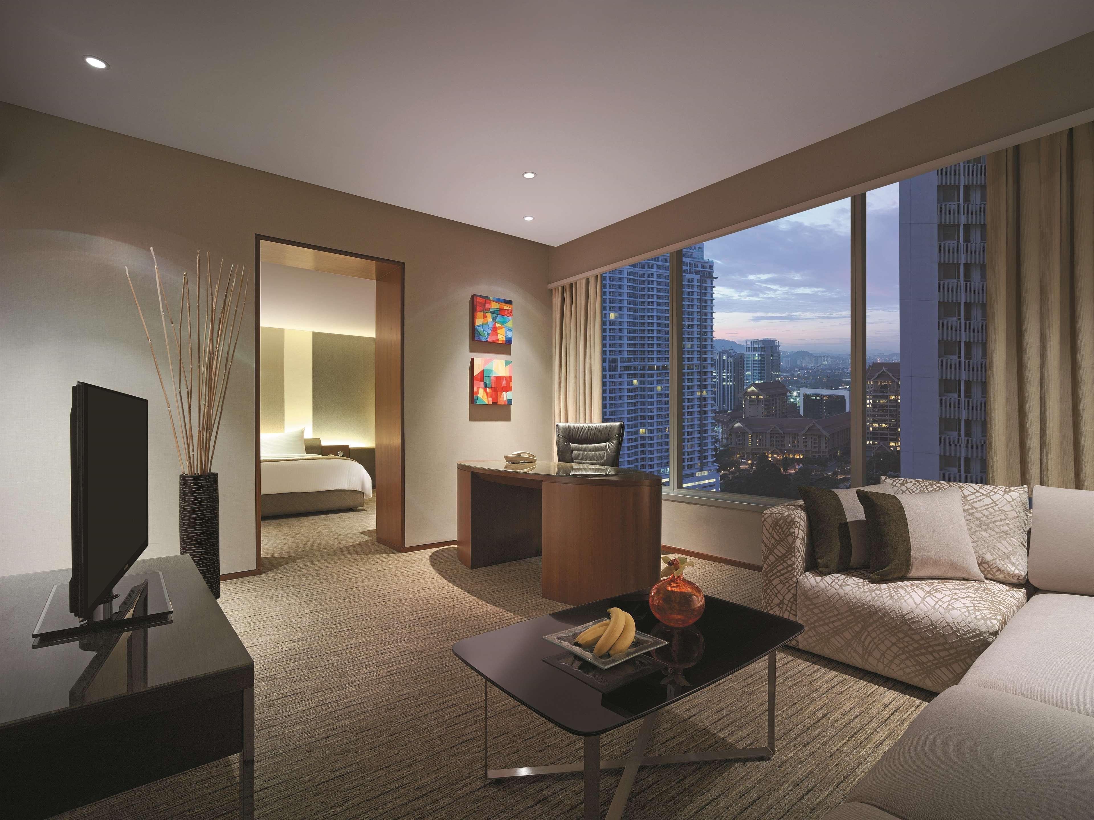 Traders Hotel, קואלה לומפור מראה חיצוני תמונה The photo shows a modern hotel room or living space. It features a comfortable-looking sofa situated next to a sleek coffee table. On the table, there are some fruits and decorative items. A flat-screen television is mounted on a cabinet opposite the