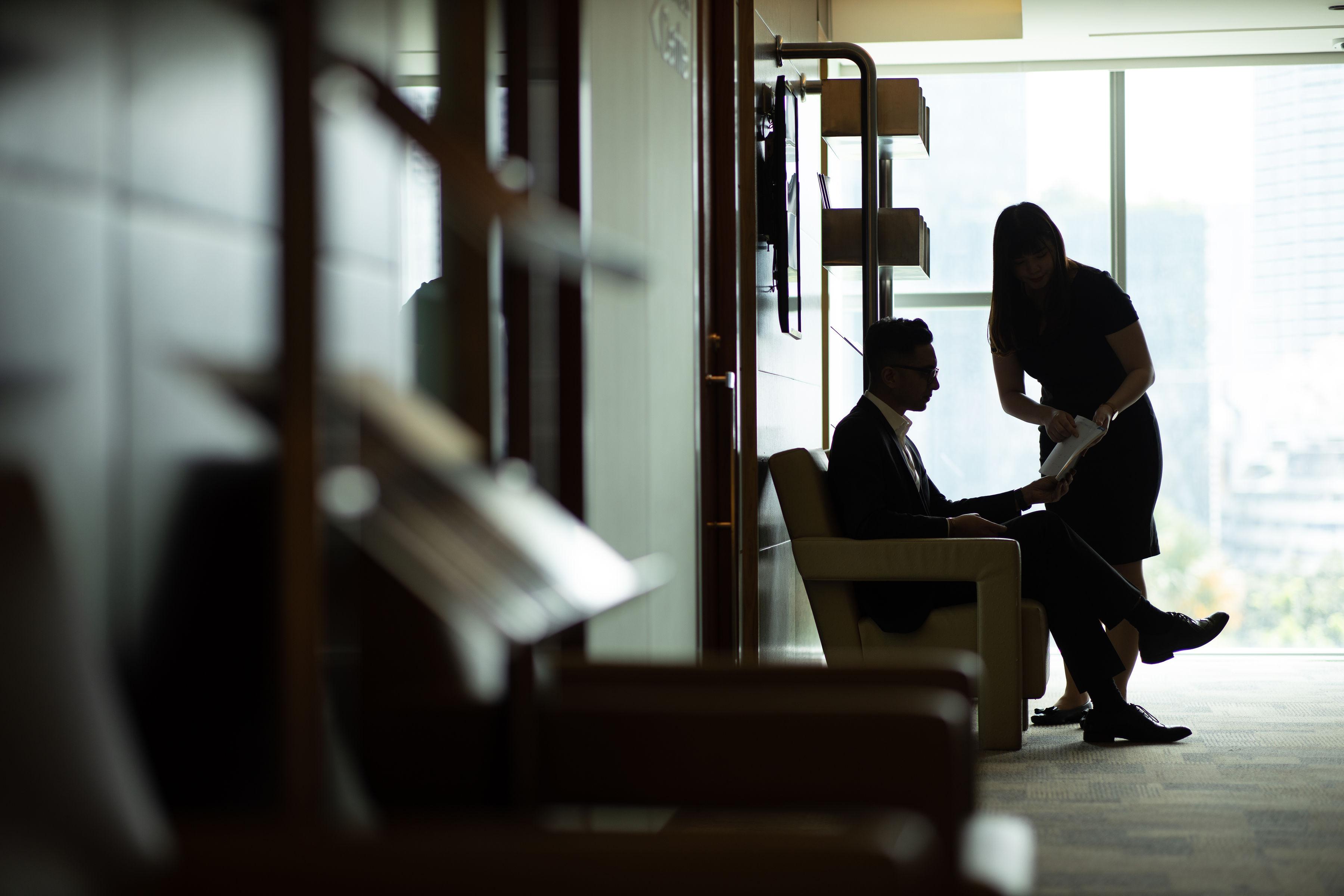 Traders Hotel, קואלה לומפור מראה חיצוני תמונה The photo shows two people in an office setting. One person, seated in a chair, is dressed in formal attire, while the other person, standing nearby, appears to be discussing something with them, possibly looking at documents. The scene is backlit, c
