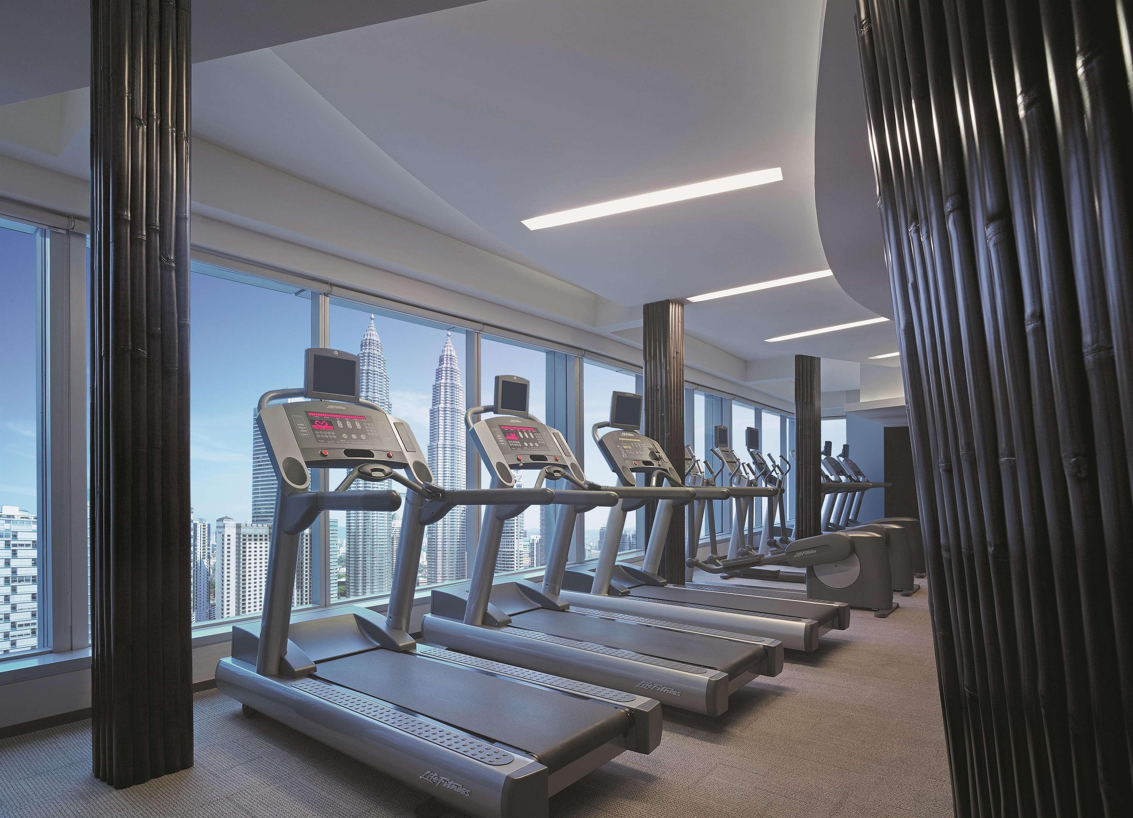 Traders Hotel, קואלה לומפור מראה חיצוני תמונה The photo shows a modern gym or fitness center. In the foreground, there are several treadmills lined up in a row. They are facing large windows that offer a view of a cityscape, featuring tall buildings and skyscrapers. The interior has a sleek desi
