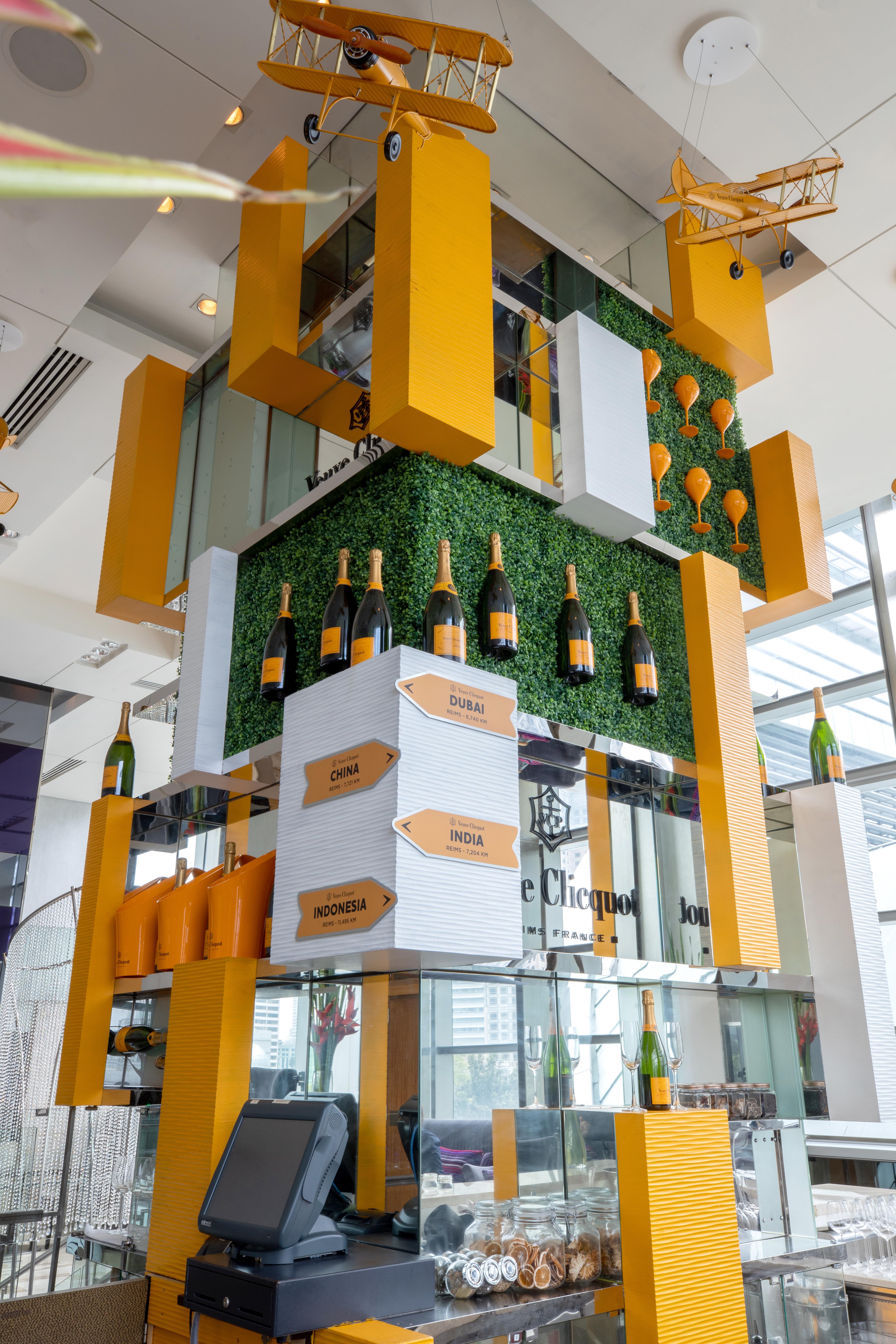 Traders Hotel, קואלה לומפור מראה חיצוני תמונה The photo depicts a stylish display featuring bottles of champagne arranged prominently on a multi-layered structure. The background includes artificial greenery, adding a touch of nature to the design. There are orange and white geometric shapes, wi