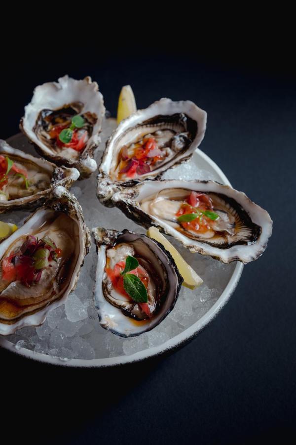 Traders Hotel, קואלה לומפור מראה חיצוני תמונה The photo shows a platter of freshly shucked oysters served on a bed of ice. Each oyster shell is filled with the tender oyster meat and topped with a vibrant mix of diced vegetables, which may include tomatoes and herbs. The oysters are accompanied 
