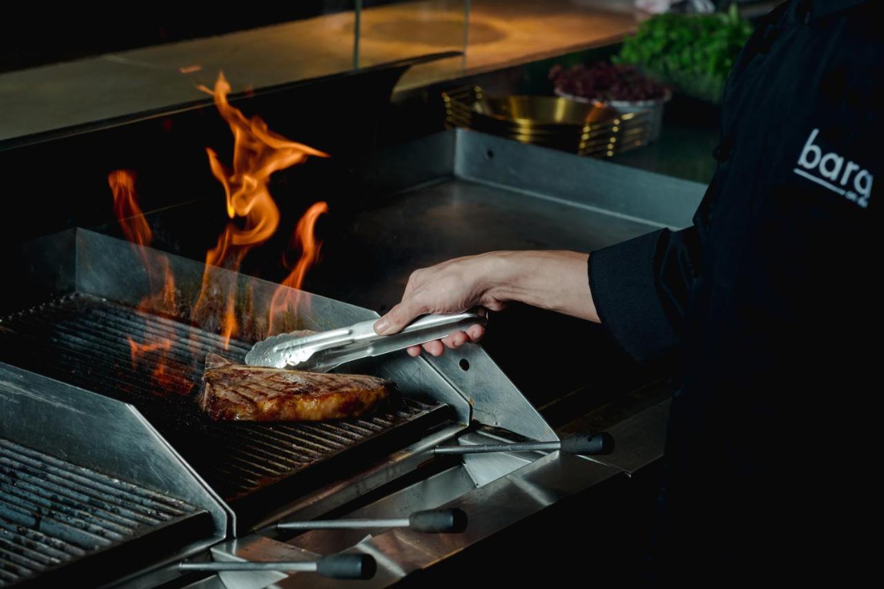 Traders Hotel, קואלה לומפור מראה חיצוני תמונה The photo shows a chef grilling a steak on a barbecue grill. There are flames rising from the grill, indicating that the steak is being cooked over high heat. The chef is using tongs to handle the steak, and the setting has a professional kitchen atm
