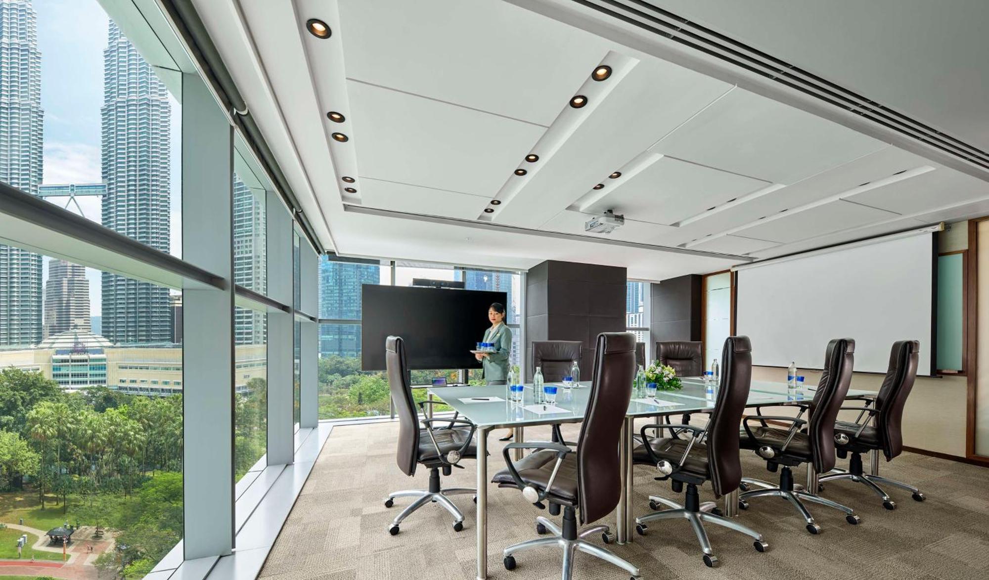 Traders Hotel, קואלה לומפור מראה חיצוני תמונה The photo shows a modern conference room with a large glass table and several ergonomic chairs arranged around it. The room features large windows that provide a view of a cityscape with tall buildings and greenery outside. There's a presentation scr