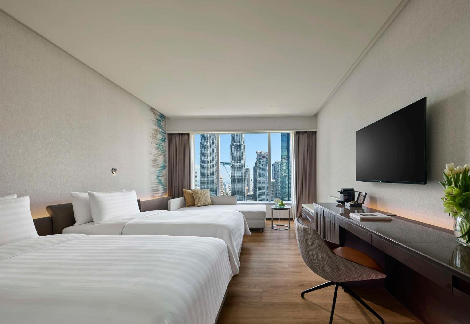 Traders Hotel, קואלה לומפור מראה חיצוני תמונה The photo shows a modern hotel room with two beds that have white linens. There is a large window with a view of city skyscrapers, allowing natural light to fill the space. A flat-screen TV is mounted on the wall, and there’s a sleek wooden desk with
