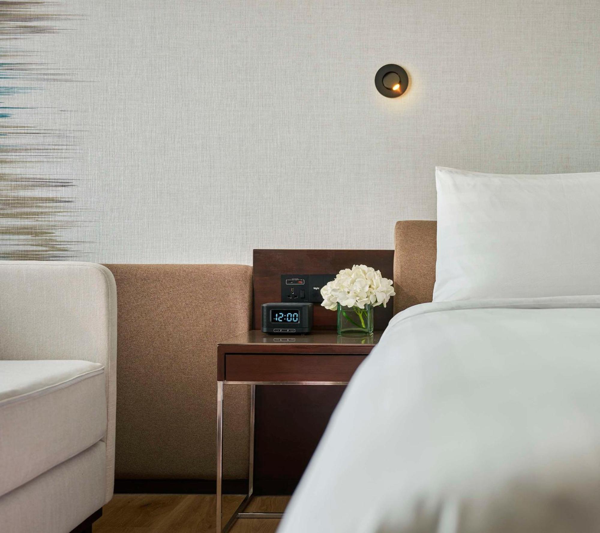 Traders Hotel, קואלה לומפור מראה חיצוני תמונה The photo shows a cozy hotel room corner featuring a bed with white bedding. Next to the bed is a small wooden nightstand that has a digital clock displaying the time. On the nightstand, there is a small bouquet of white flowers, and beside it, you c