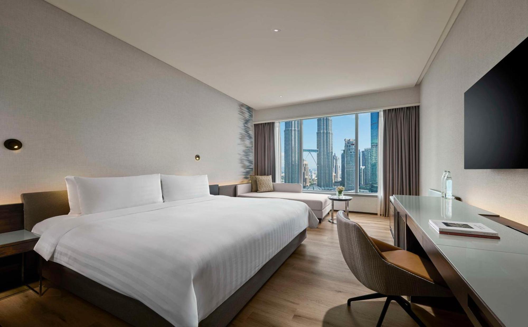Traders Hotel, קואלה לומפור מראה חיצוני תמונה The photo shows a modern hotel room featuring a large bed with white linens. There is a comfortable seating area by the window, which provides a view of tall buildings outside. The room is well-lit and has a minimalistic design, with neutral-colored 