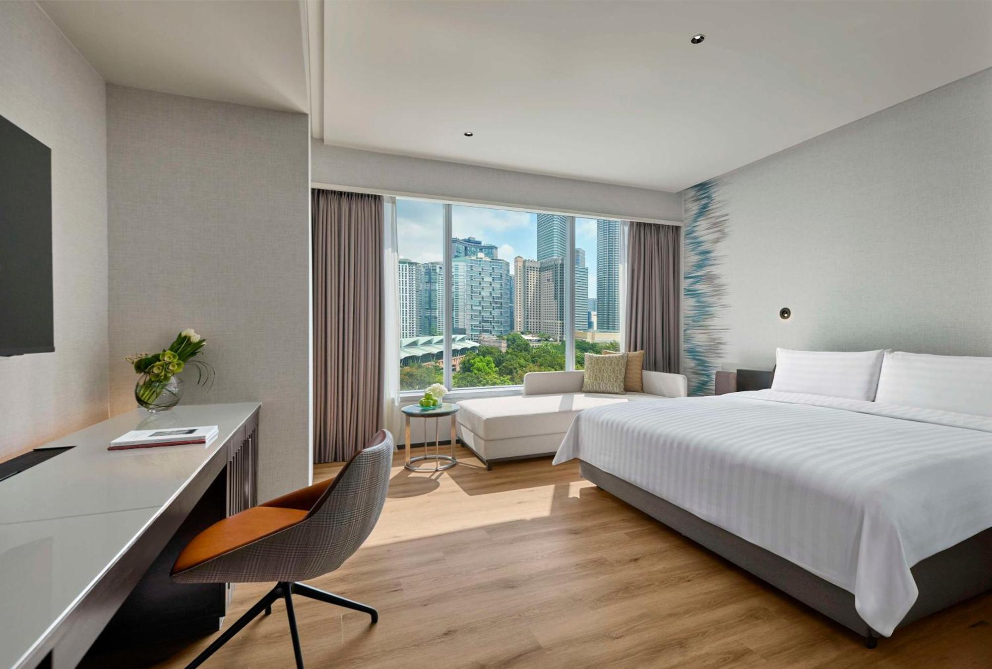 Traders Hotel, קואלה לומפור מראה חיצוני תמונה The photo shows a modern hotel room featuring a large bed with white bedding. There is a stylish chair and desk on one side, and a cozy lounge area with a sofa near the window. Large windows provide a view of a city skyline, allowing natural light to