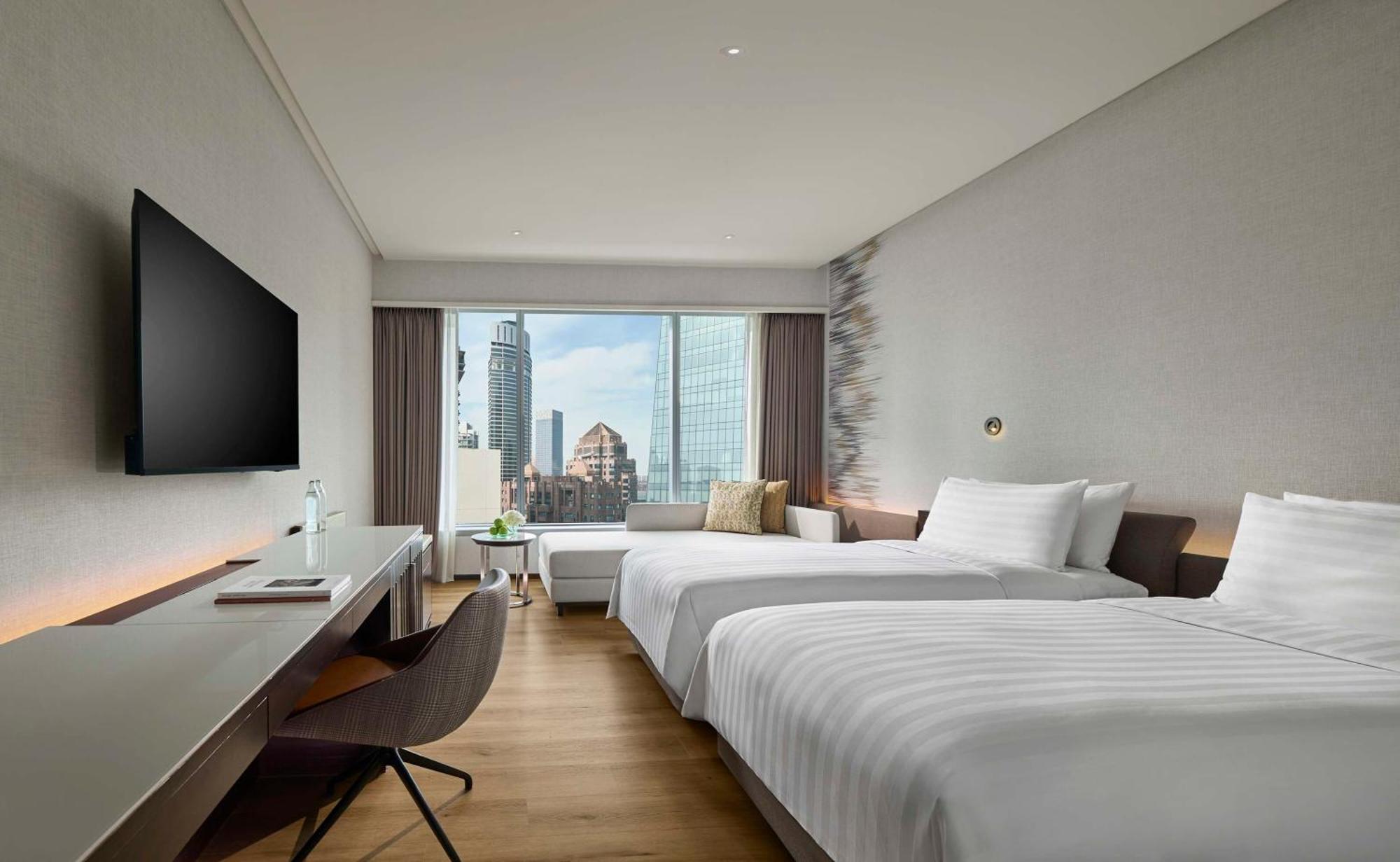 Traders Hotel, קואלה לומפור מראה חיצוני תמונה The photo shows a modern hotel room featuring two single beds with crisp white linens. There is a large window providing a panoramic view of the city skyline. In front of the window, there is a small seating area with a cushion. A flat-screen TV is m