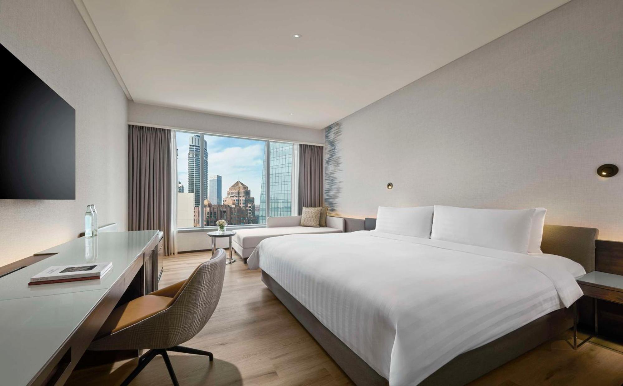 Traders Hotel, קואלה לומפור מראה חיצוני תמונה The photo shows a modern hotel room featuring a large bed with crisp white linens. There's a window with a view of a city skyline, allowing natural light to fill the space. The room includes a small seating area near the window, and a sleek desk with