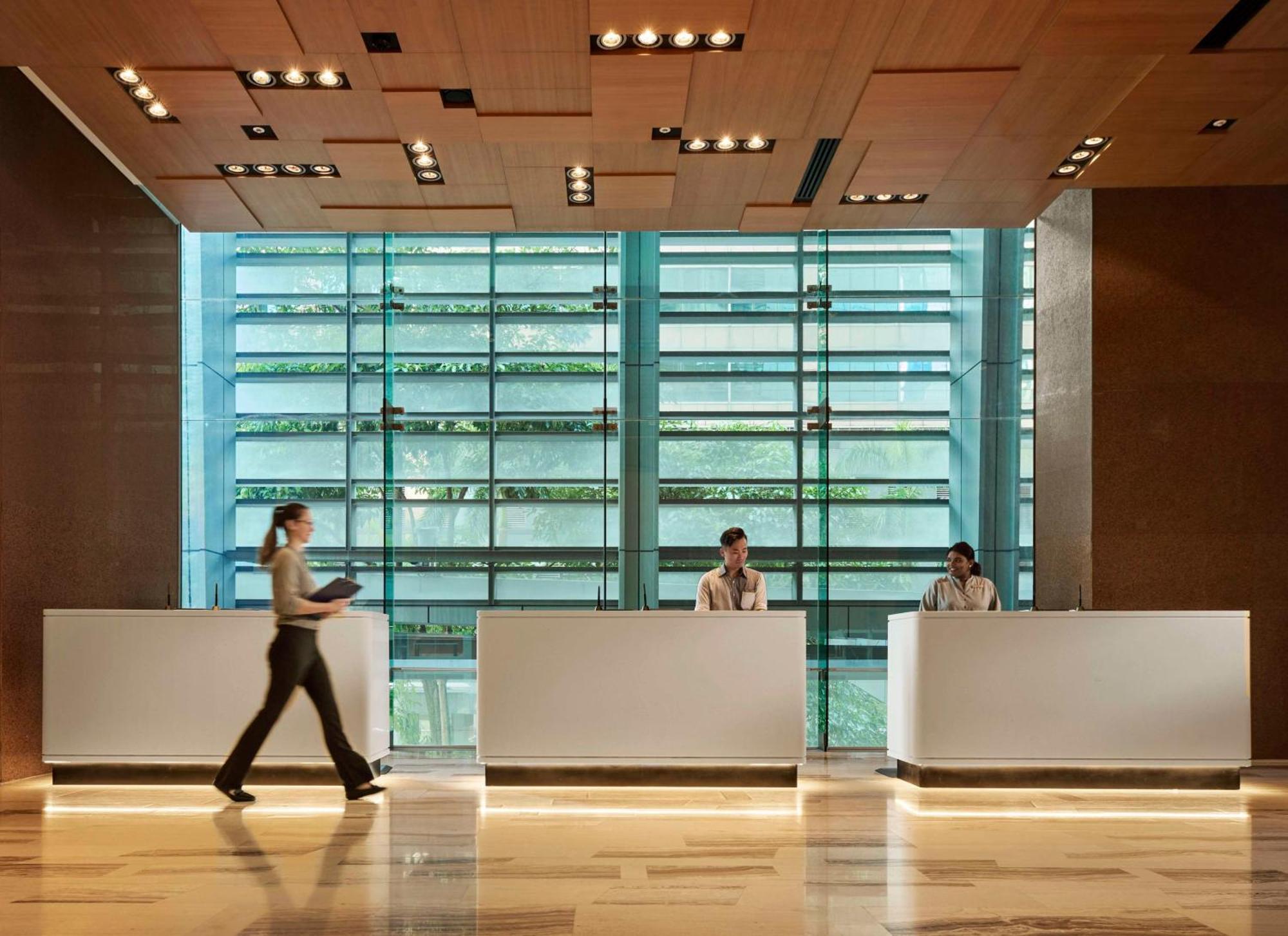 Traders Hotel, קואלה לומפור מראה חיצוני תמונה The photo shows a modern reception area featuring three white reception desks in a spacious, well-lit environment. Behind the desks is a large glass wall with horizontal slats that allows natural light to filter through while maintaining some privacy