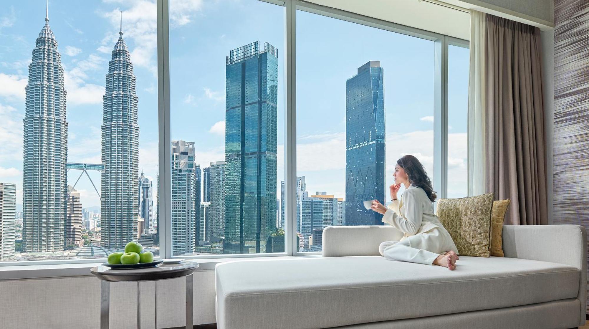 Traders Hotel, קואלה לומפור מראה חיצוני תמונה The photo shows a woman sitting on a comfortable couch, gazing out of a large window. She is dressed in cozy, light-colored clothing and holding a cup, possibly enjoying a beverage. Outside the window, there is a panoramic view of a modern cityscape,