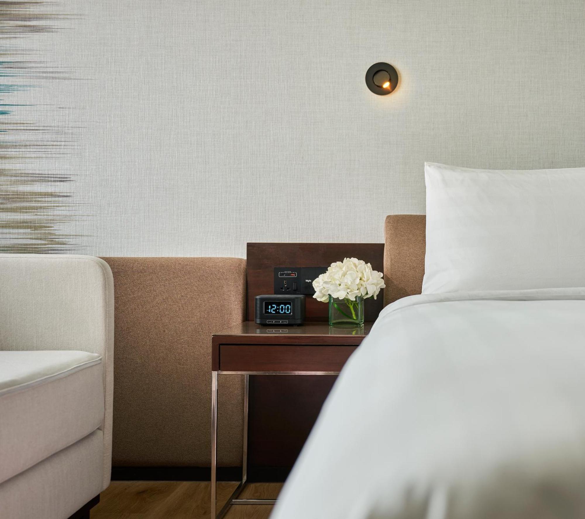 Traders Hotel, קואלה לומפור מראה חיצוני תמונה The photo shows a cozy bedroom setting. To the right, there's a neatly made bed with white bedding. Next to the bed, there's a wooden bedside table with a small vase of white flowers on it. On the table, there is also a digital clock displaying the t