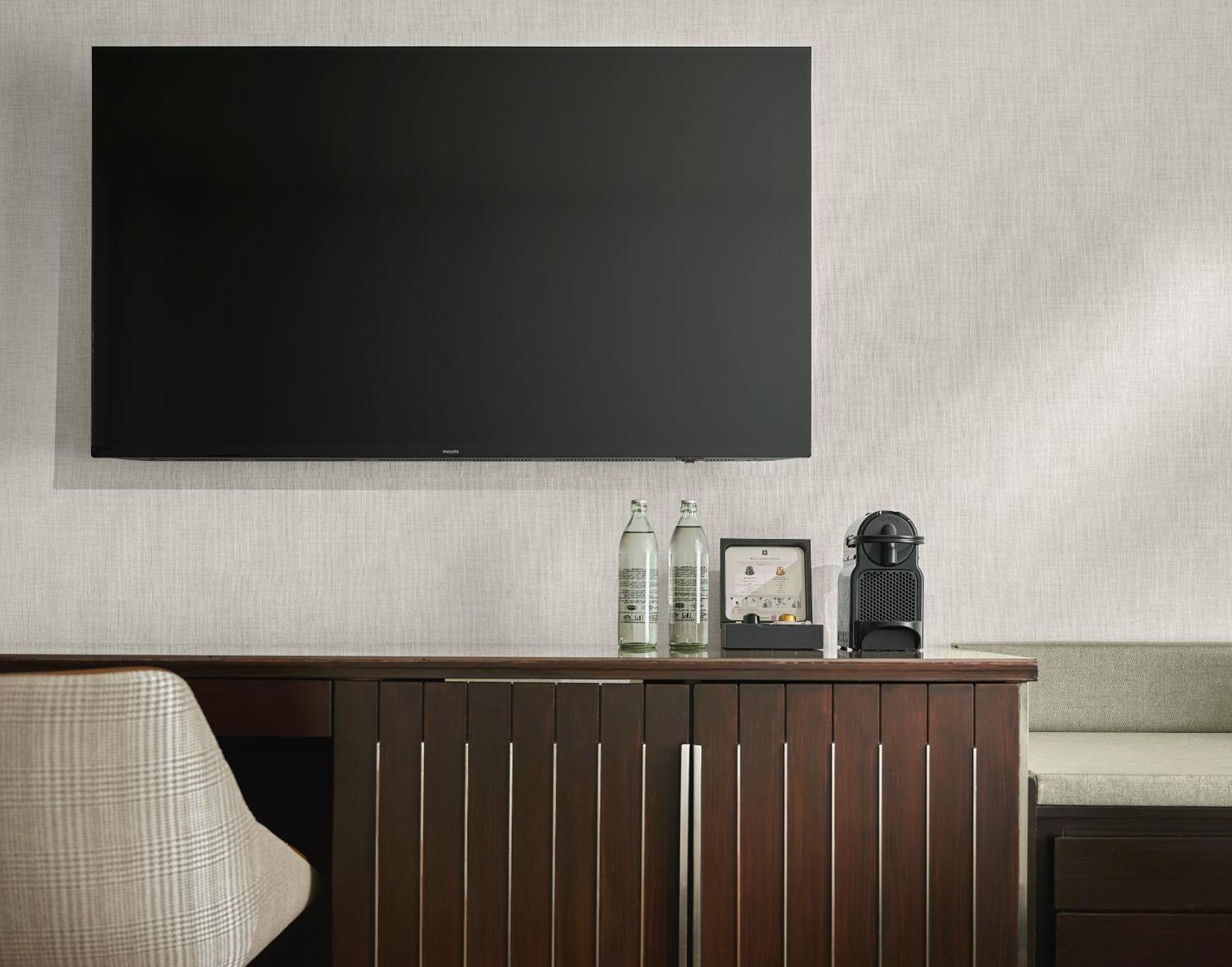 Traders Hotel, קואלה לומפור מראה חיצוני תמונה The photo shows a contemporary room setting with a flat-screen television mounted on the wall. Below the TV, there is a wooden cabinet or sideboard with closed storage compartments. On top of the cabinet, there are two bottles of water, a small decor