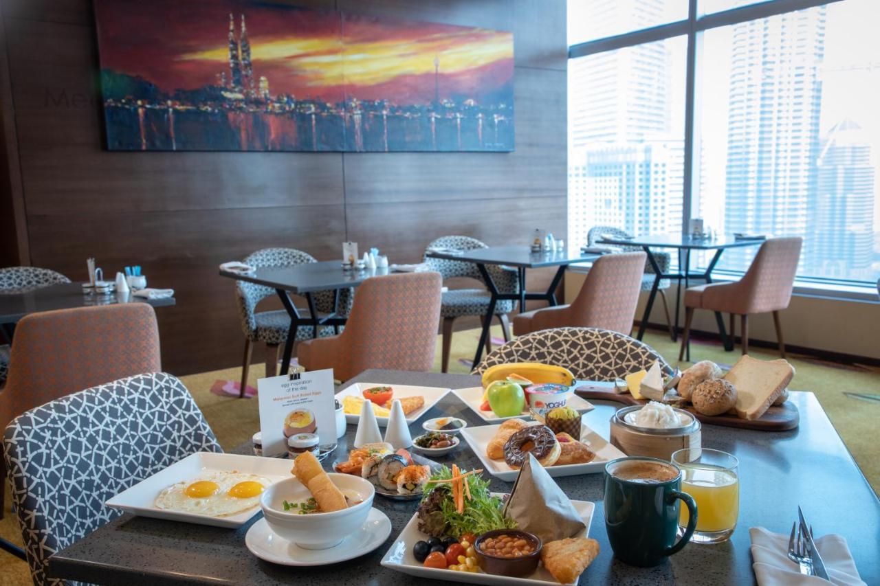 Traders Hotel, קואלה לומפור מראה חיצוני תמונה The photo shows a dining area with a modern decor featuring a colorful painting of a city skyline at sunset. There are several tables with chairs, and on one of the tables, there's a variety of breakfast items. The spread includes plates of food with